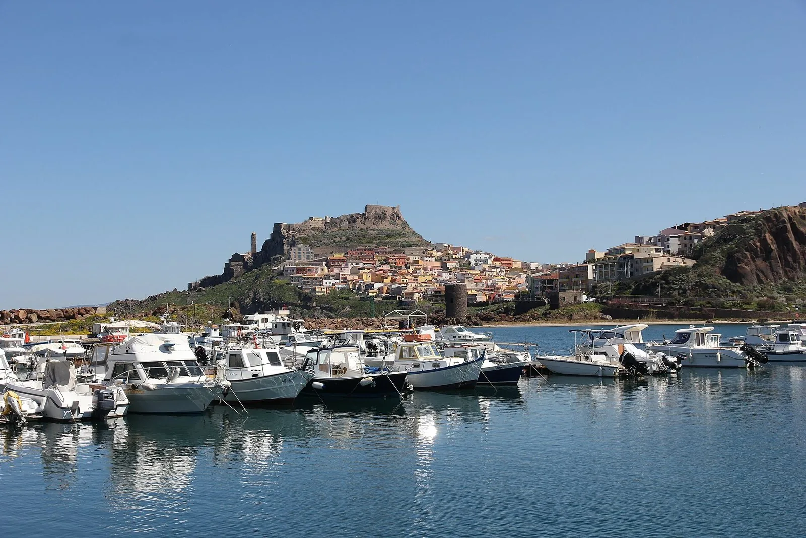 Castelsardo visto dal porticciolo: barche ormeggiate in primo piano, case colorate che salgono sul promontorio fino al castello e al campanile