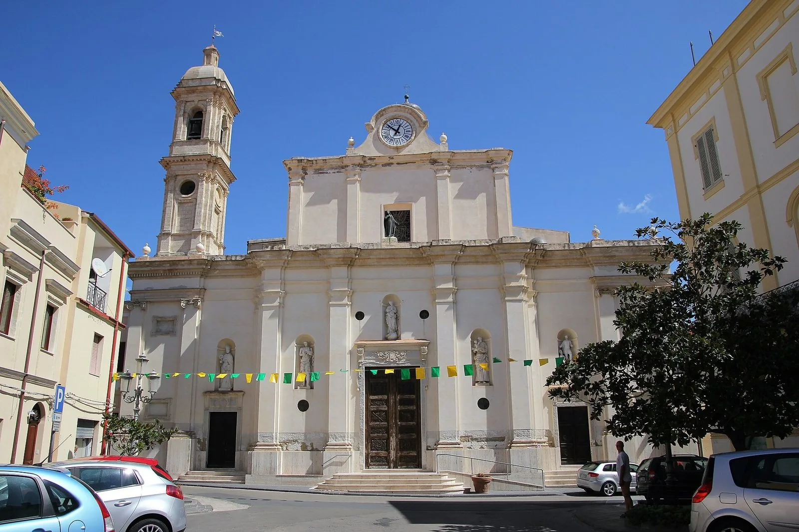 La facciata neoclassica chiara della chiesa parrocchiale di San Pantaleo a Sorso, con campanile, statue nelle nicchie e bandierine colorate, sotto un cielo azzurro