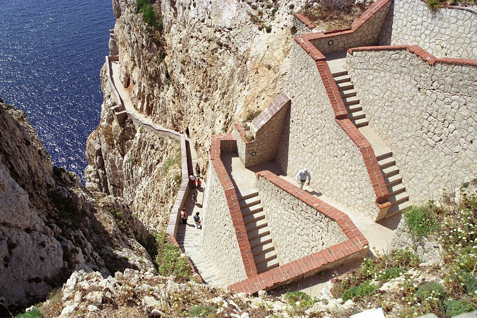 L'Escala del Cabirol vista dall'alto: rampe di scale in pietra con bordi in cotto che scendono lungo la parete di Capo Caccia verso il mare