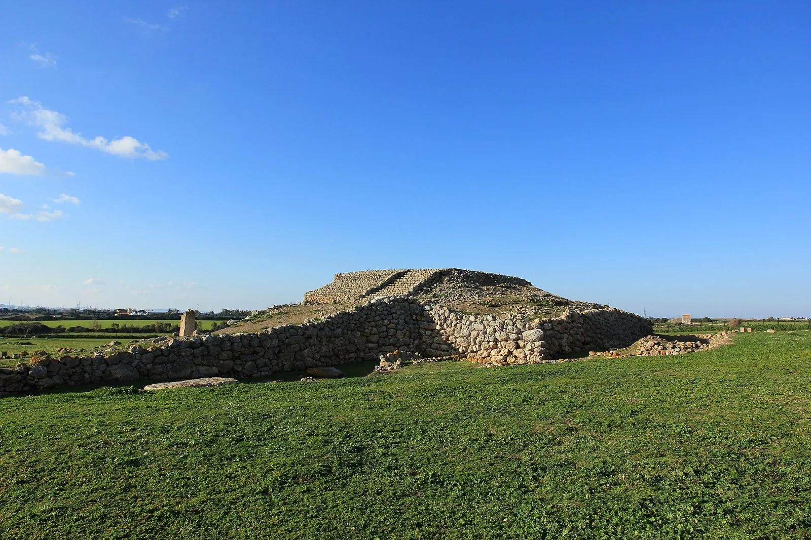 L'altare prenuragico di Monte d'Accoddi: la grande terrazza in pietra con la rampa d'accesso, un menhir sulla sinistra e un prato verde in primo piano sotto il cielo azzurro