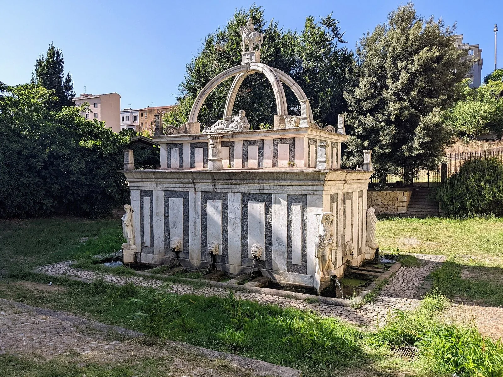 La Fontana di Rosello a Sassari, in marmo chiaro e pietra scura, con le statue delle stagioni agli angoli e la statua equestre di San Gavino sopra due archi incrociati, circondata da prato e alberi